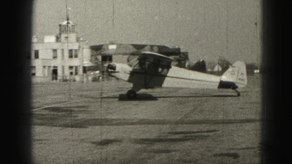 Small propeller plane on a Harrisburg airport tarmac in 1975 home-movie footage, with a low building behind it.