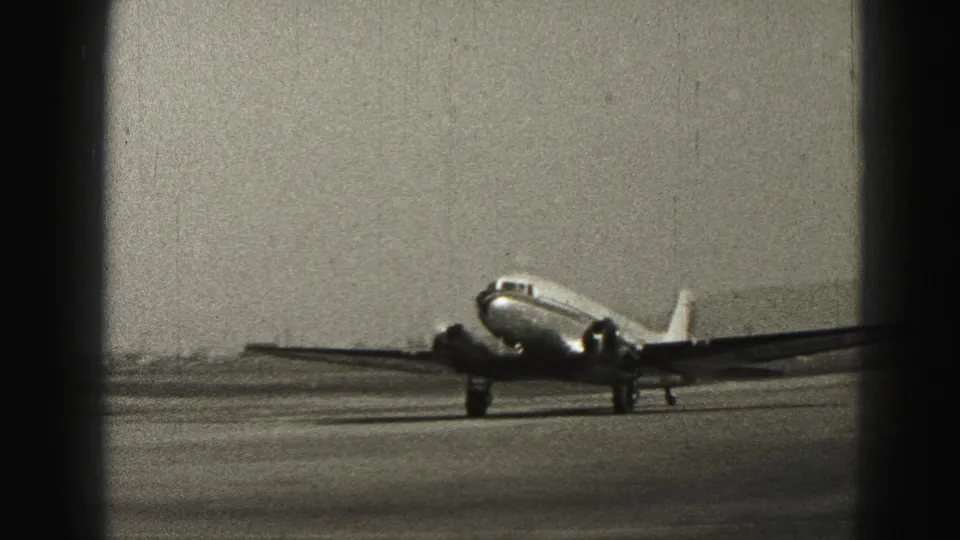 1975 Harrisburg home-movie frame showing a small propeller passenger aircraft taxiing across airport pavement.