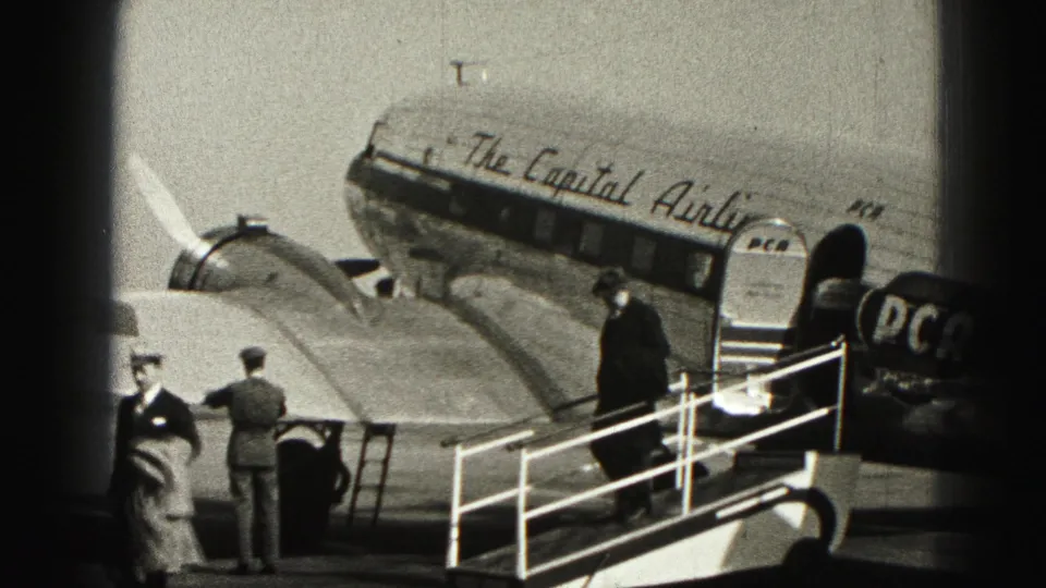 1975 Harrisburg home-movie frame showing men near mobile stairs and a Capital Airlines propeller aircraft on the ramp.