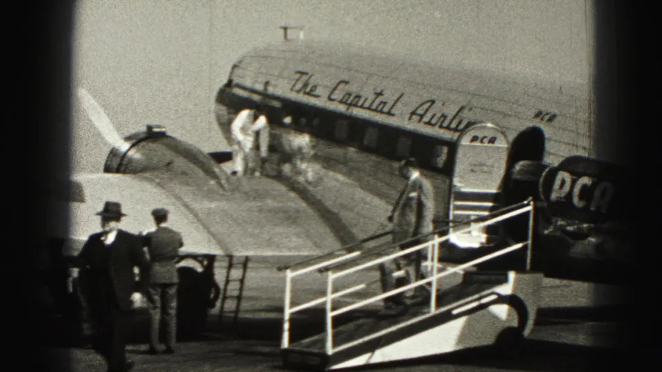 Home-movie frame of men on an airport ramp near a Capital Airlines plane, with boarding stairs and crew visible.