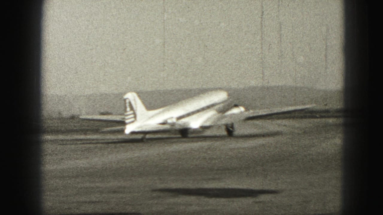 1975 Harrisburg home-movie frame showing a propeller airplane on the runway, seen through dark film-edge shadows.