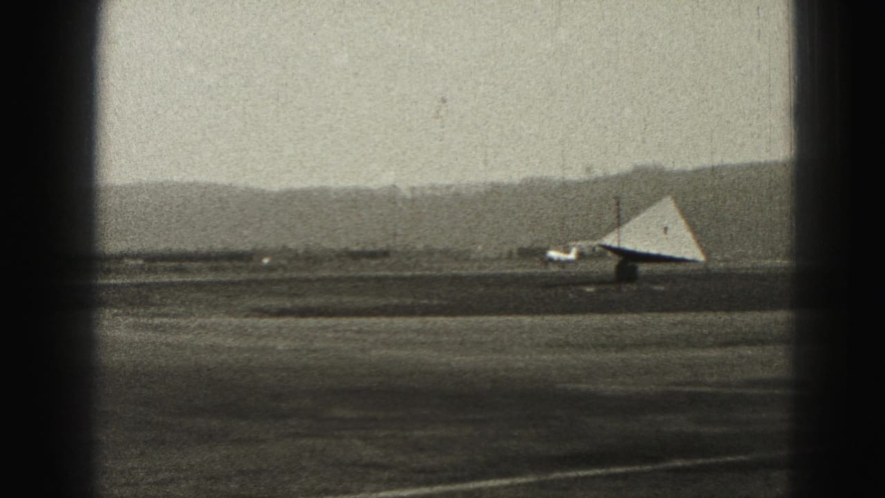1975 8mm frame from Harrisburg showing a small aircraft taxiing across an airfield beneath a hazy sky and distant hills.