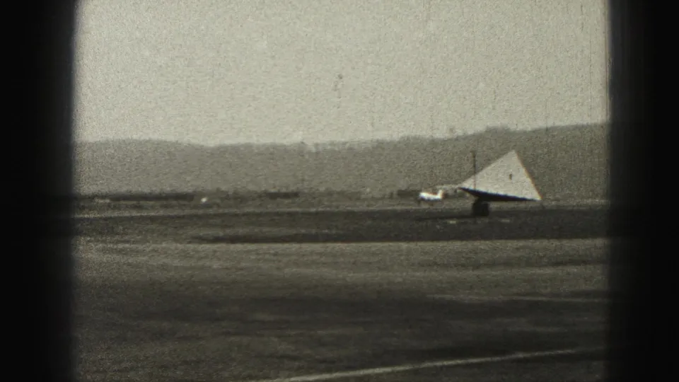1975 8mm frame from Harrisburg showing a small aircraft taxiing across an airfield beneath a hazy sky and distant hills.