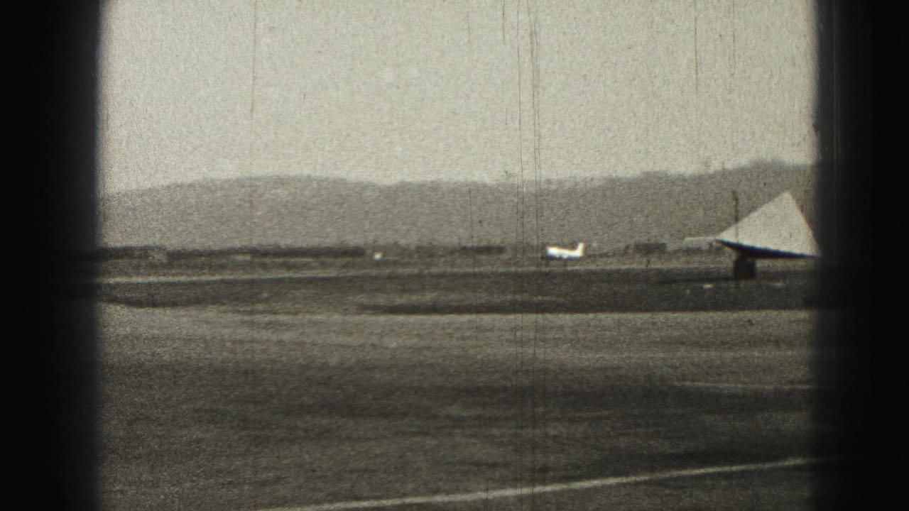 1975 Harrisburg airfield view with a small airplane crossing the tarmac near a triangular structure and distant hills.