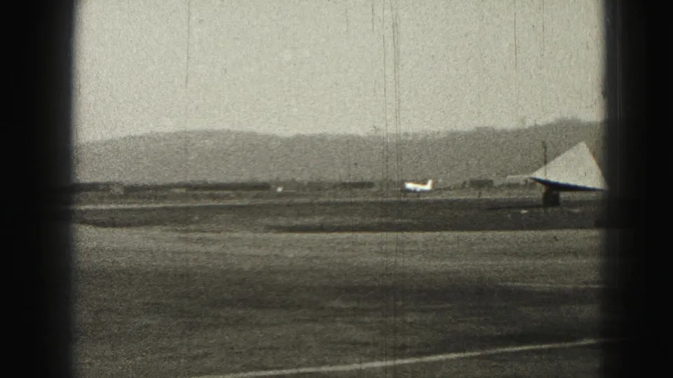 1975 Harrisburg airfield view with a small airplane crossing the tarmac near a triangular structure and distant hills.