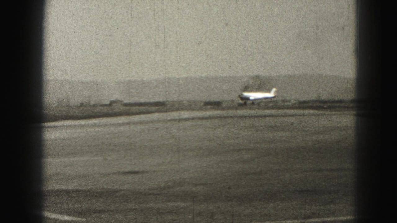 1975 Harrisburg-area 8mm frame of a small propeller plane just above the runway at a rural airport.
