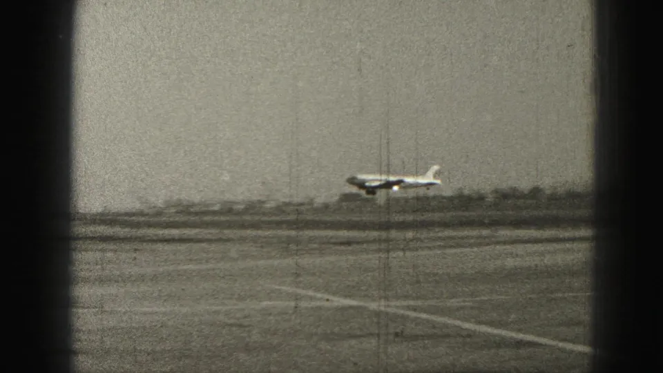 1975 Harrisburg-area 8mm frame of a small plane flying low over an airfield with runway lines and haze behind it.