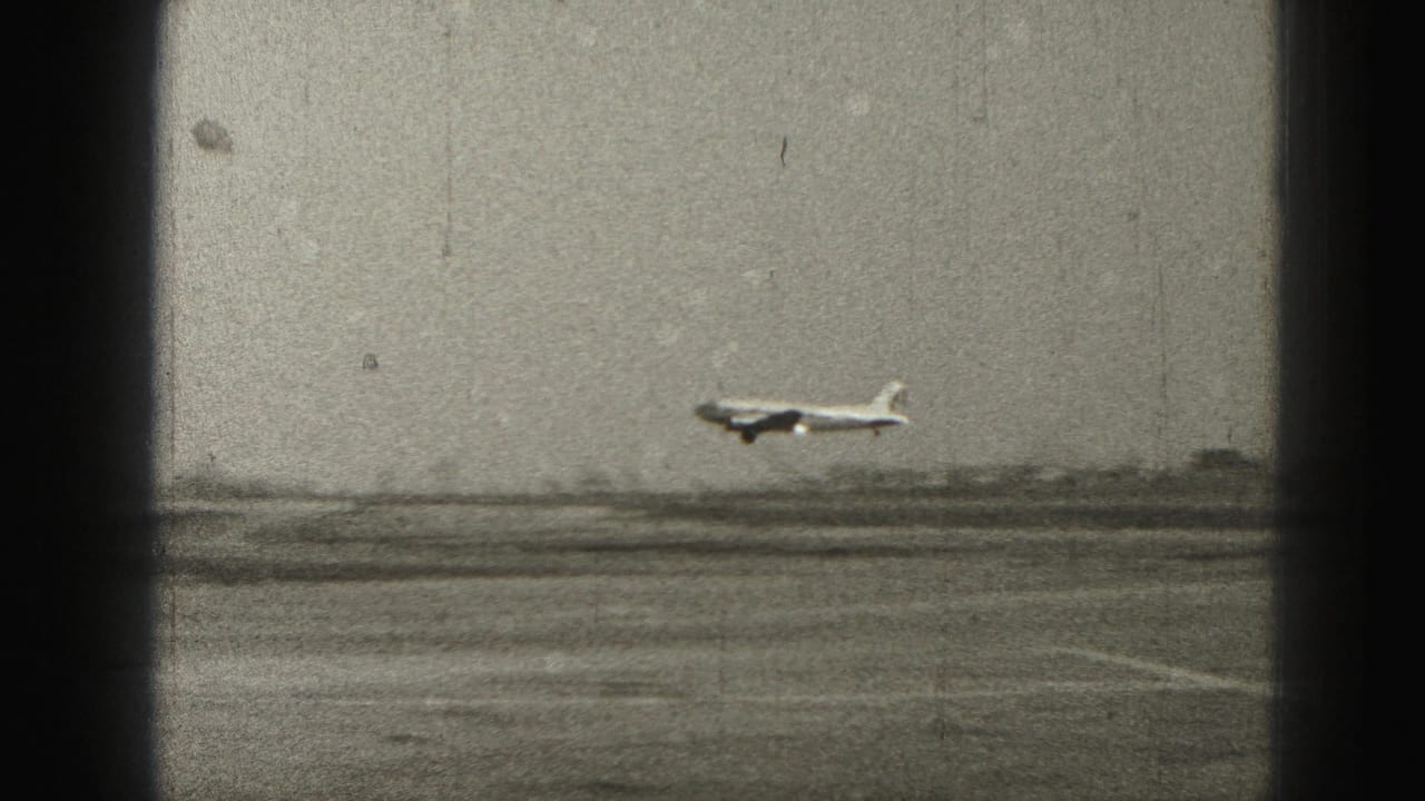 1975 Harrisburg-area 8mm frame showing a large light-colored airplane flying low over open land behind a dark window frame.
