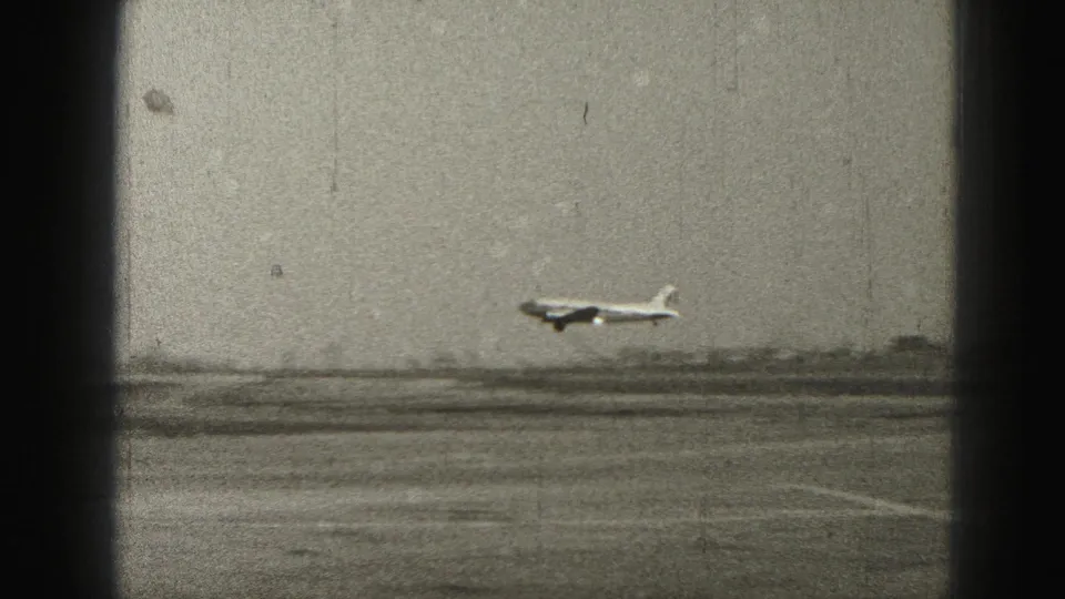 1975 Harrisburg-area 8mm frame showing a large light-colored airplane flying low over open land behind a dark window frame.