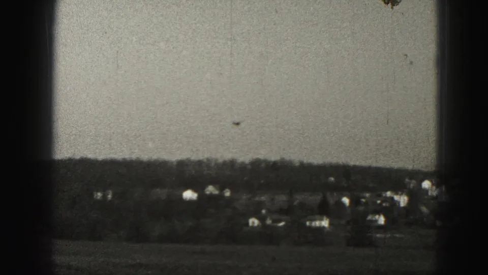 1975 Harrisburg-area 8mm frame showing distant suburban houses and a treeline across open ground, with no people visible.