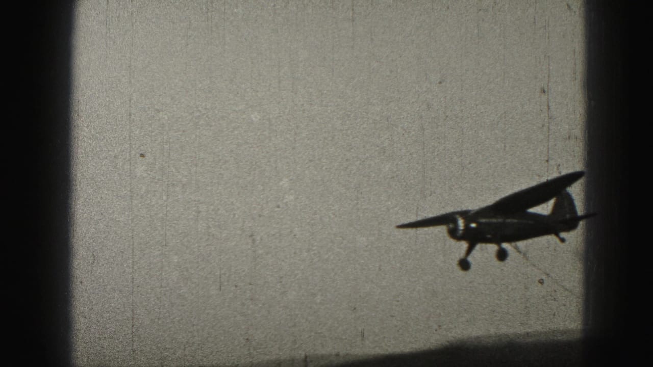 A small propeller aircraft crosses low in the frame, with landing gear visible against the pale 1975 8mm sky.