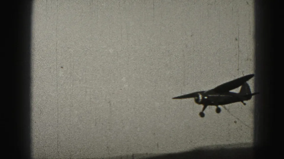 A small propeller aircraft crosses low in the frame, with landing gear visible against the pale 1975 8mm sky.