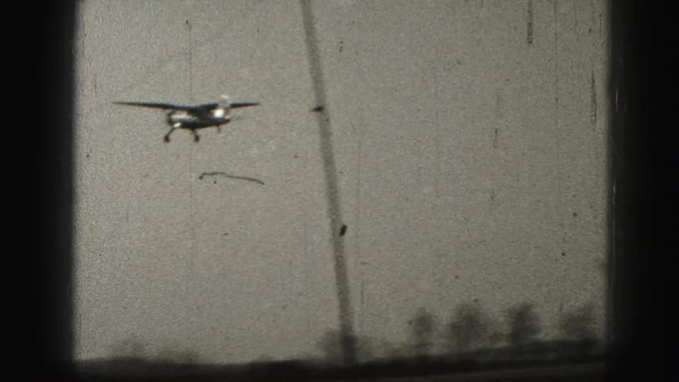 The small airplane appears head-on or quartering toward camera above a low landscape, with 8mm grain and scratches visible.
