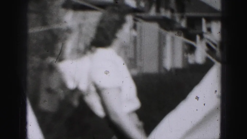 Blurred 1975 Harrisburg home-movie frame of a woman in a light dress standing outdoors near branches and a porch-like structure.