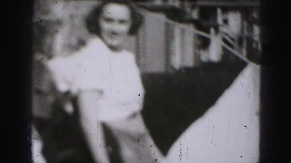 Soft, grainy 1975 Harrisburg home-movie frame of a woman facing the camera outdoors near branches and a building or porch detail.