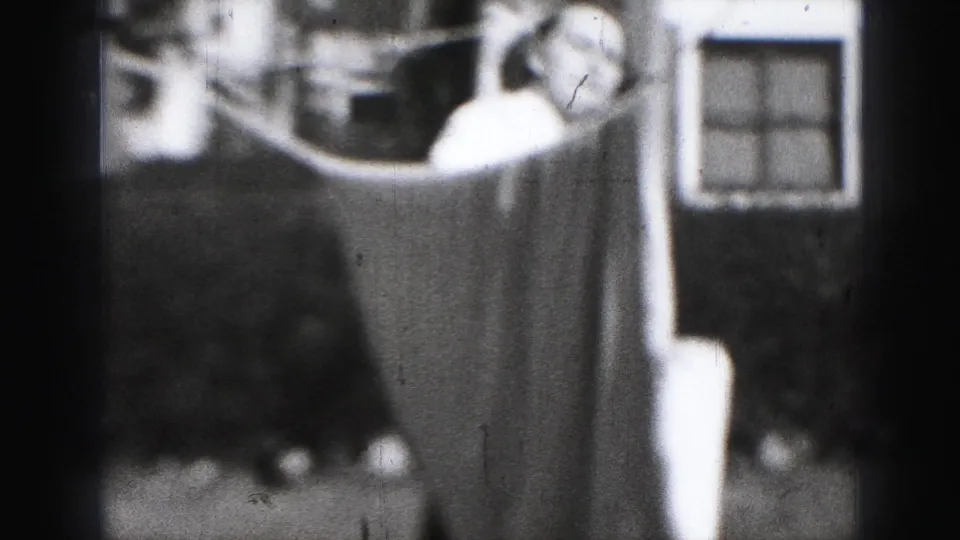 A woman looks over or around a sheet on a clothesline, with a window or door frame visible in the background.