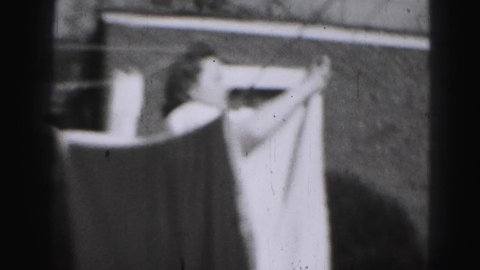 The woman bends or reaches beside a large sheet on the clothesline, framed by soft 8mm film texture.