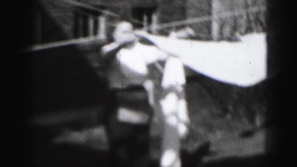 1975 Harrisburg home-movie frame of a woman securing a large light sheet on a yard clothesline.