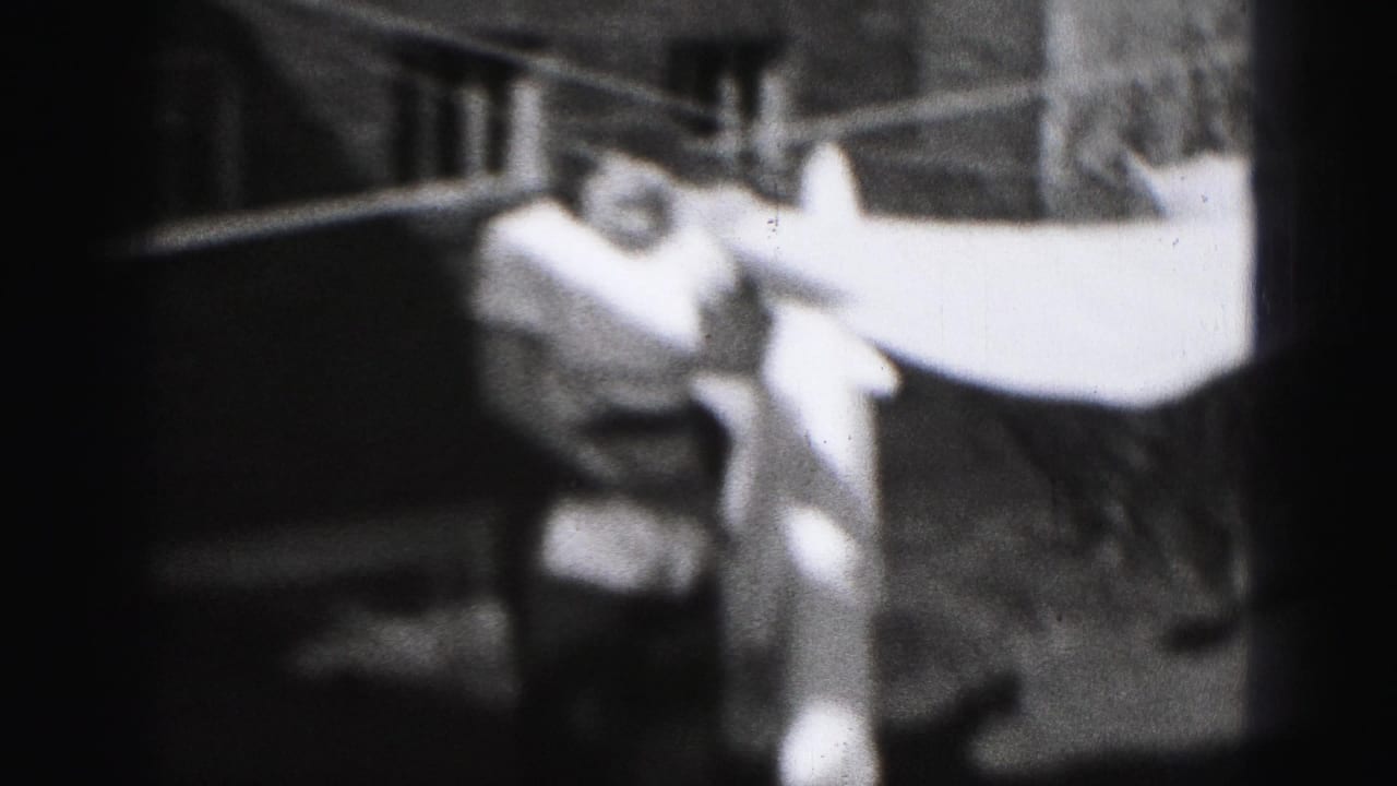 In 1975 Harrisburg home-movie footage, a woman in a light dress reaches up to hang white laundry outdoors.