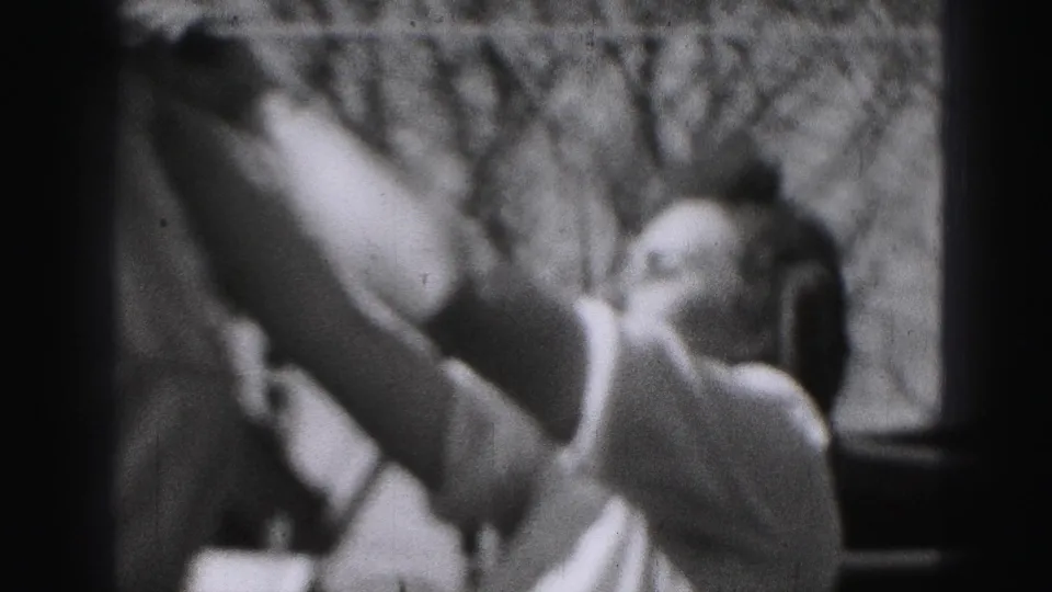 The woman appears beside a large white garment, reaching up as bare branches fill the background of the 1975 home movie.