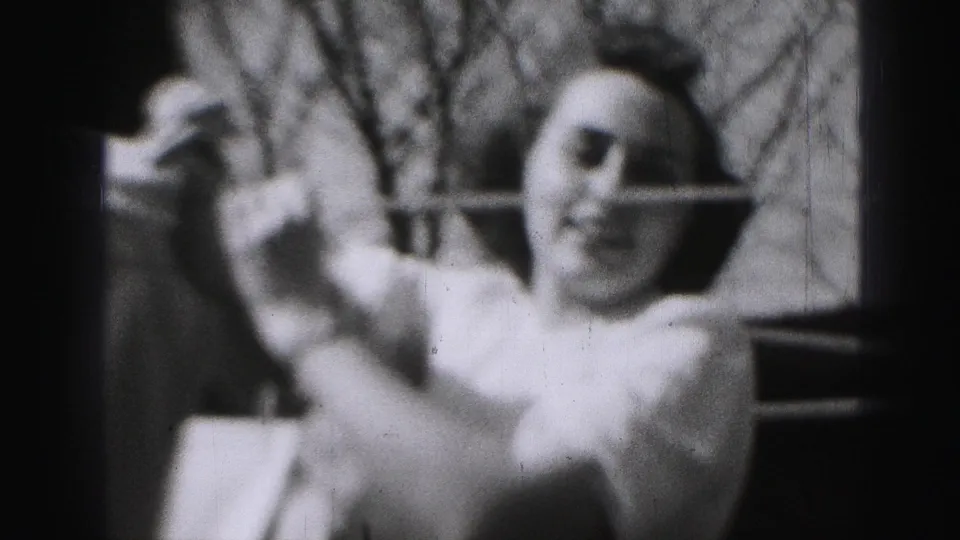In 1975 Harrisburg home-movie footage, a woman in a light shirt reaches toward a backyard clothesline with bare trees behind her.