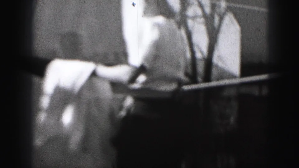 The woman leans near the clothesline and railing, with hanging fabric and bare trees visible in the 1975 Harrisburg home-movie frame.
