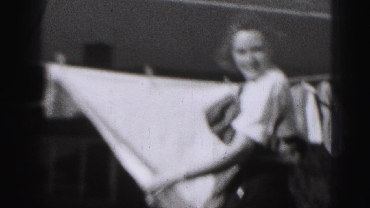 In Harrisburg 1975 home-movie footage, a woman in a white blouse and dark skirt stands by a sheet on a backyard clothesline.