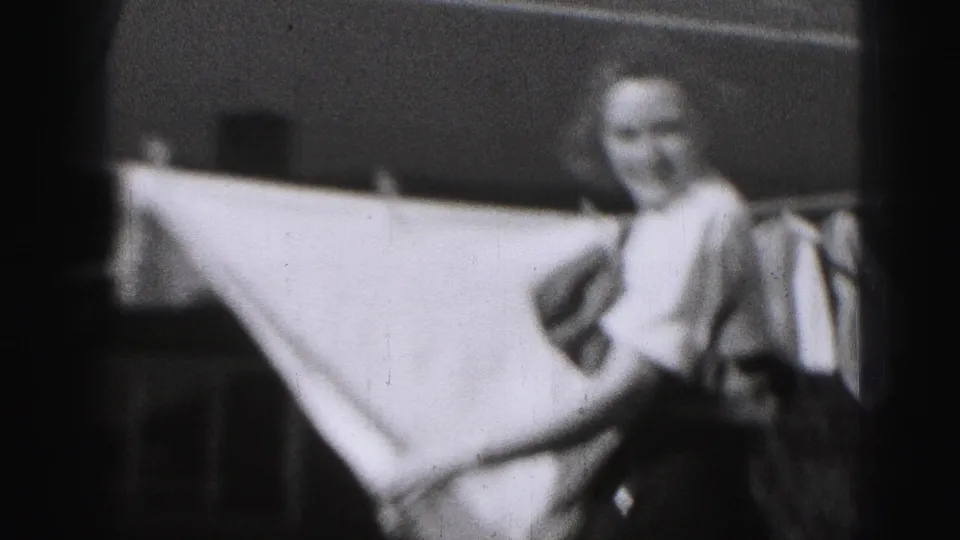 In Harrisburg 1975 home-movie footage, a woman in a white blouse and dark skirt stands by a sheet on a backyard clothesline.