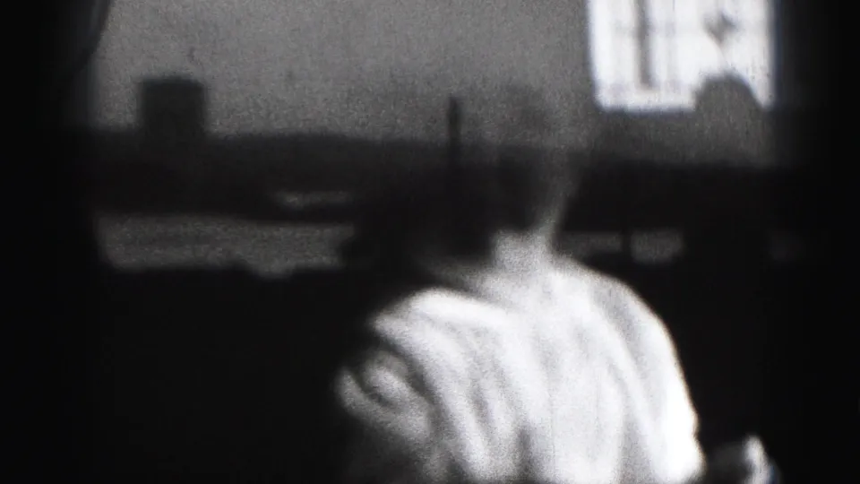 1975 8mm home-movie frame from Harrisburg showing a baby in a light outfit, softly focused against a bright interior window or screen.