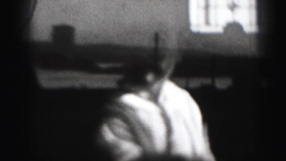 Grainy 1975 8mm frame showing the baby turned sideways in a dim Harrisburg interior, with a bright window or screen behind them.