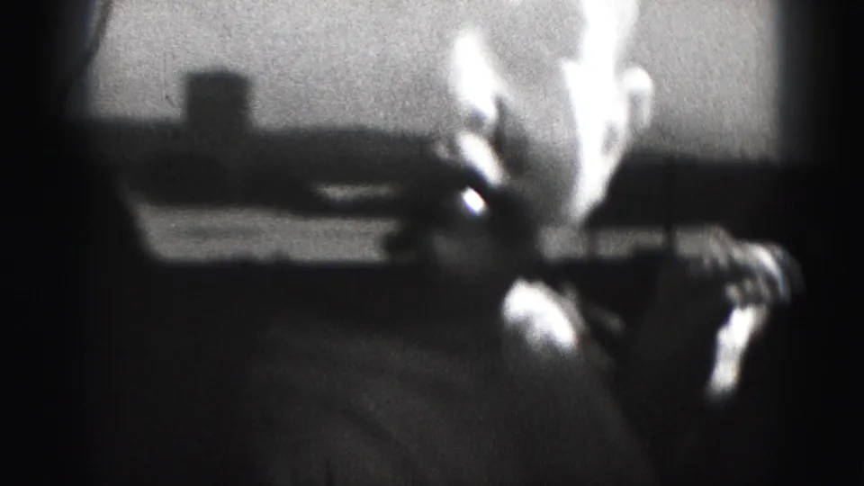Black-and-white 8mm frame from 1975 showing a close, overexposed view of a baby leaning near the camera in a dim room.