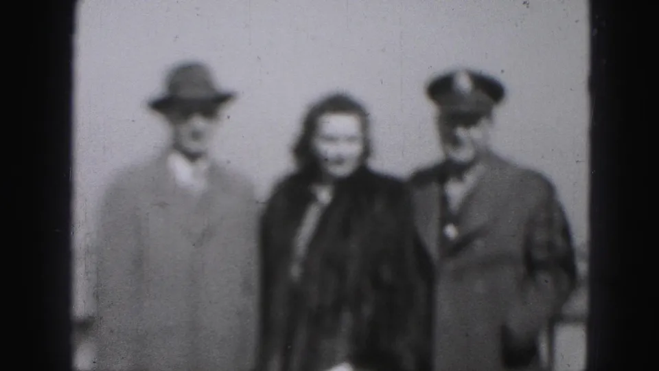 Soft black-and-white 8mm frame from Harrisburg, Pennsylvania, showing a woman in a dark fur coat between a hatted man and a uniformed man.