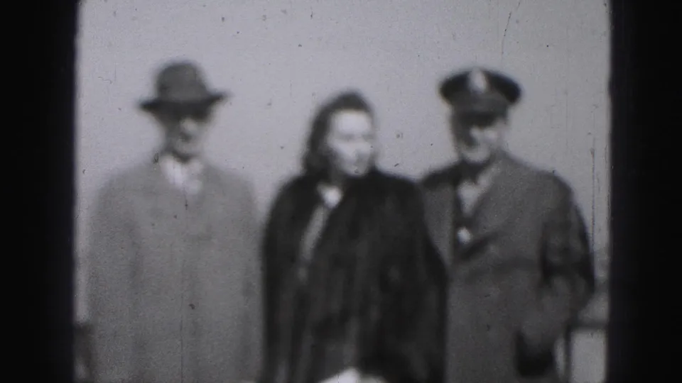 Black-and-white 8mm frame shows the group facing forward, with the uniformed man at right and a hatted civilian at left.
