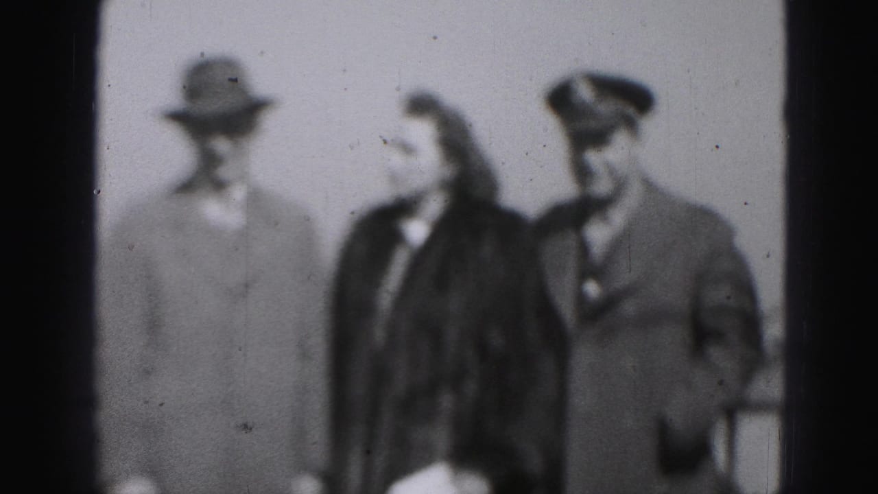 Three adults in coats and hats stand close together in soft-focus black-and-white footage.