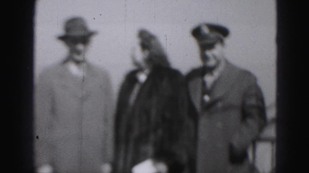 In this 1975 Harrisburg home-movie frame, a woman in a dark coat stands between two men, one in a peaked cap.