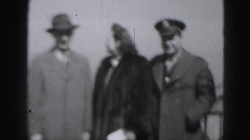 In this 1975 Harrisburg home-movie frame, a woman in a dark coat stands between two men, one in a peaked cap.