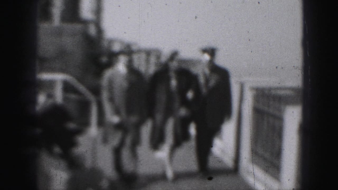 Three adults walk along a ship deck beside railings in soft black-and-white 8mm footage.