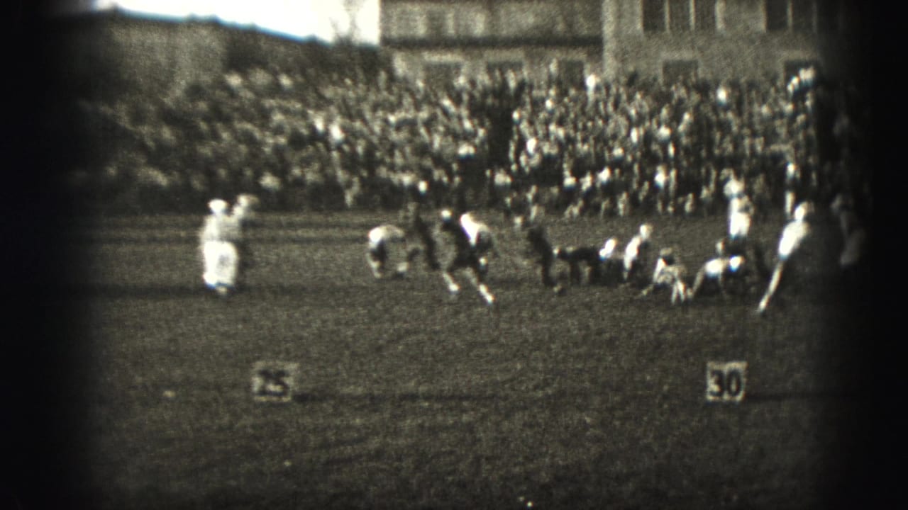 During the 1975 Harrisburg football game, players in light and dark uniforms move through a play before a large crowd.