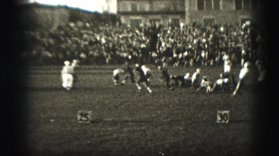 During the 1975 Harrisburg football game, players in light and dark uniforms move through a play before a large crowd.