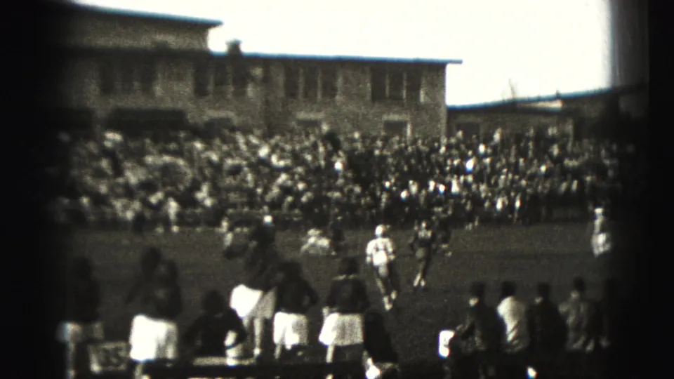 Sideline spectators or cheerleaders stand near the field as 1975 Harrisburg football players continue play before packed bleachers.
