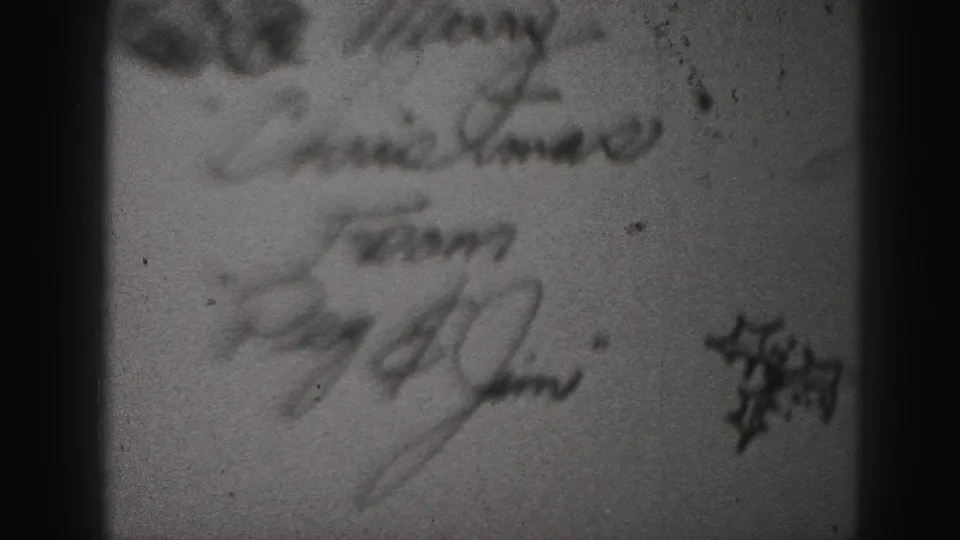 Blurred 1975 8mm view of a handwritten holiday note with signatures and a small festive-looking flourish.