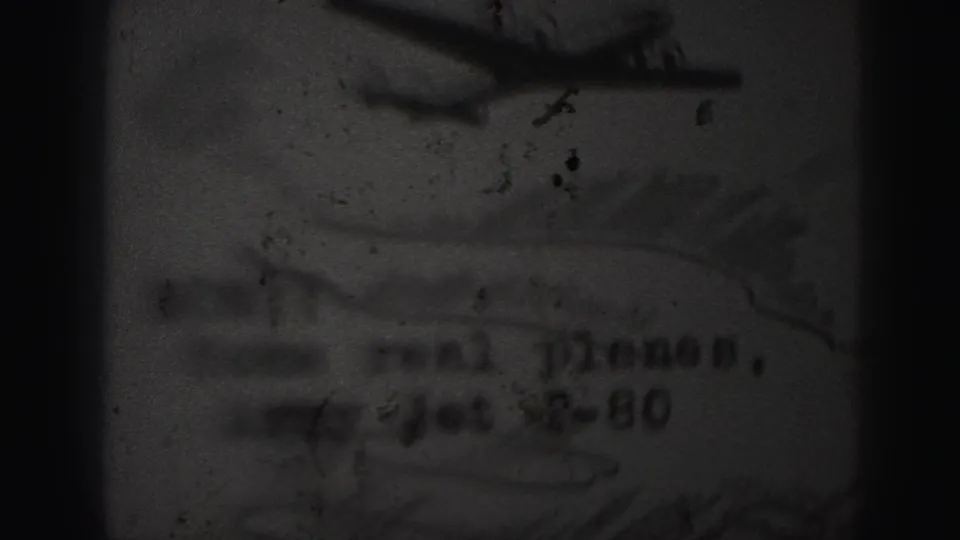 Grainy 8mm frame showing a child's-style drawing of a jet aircraft with partly readable lettering beneath.
