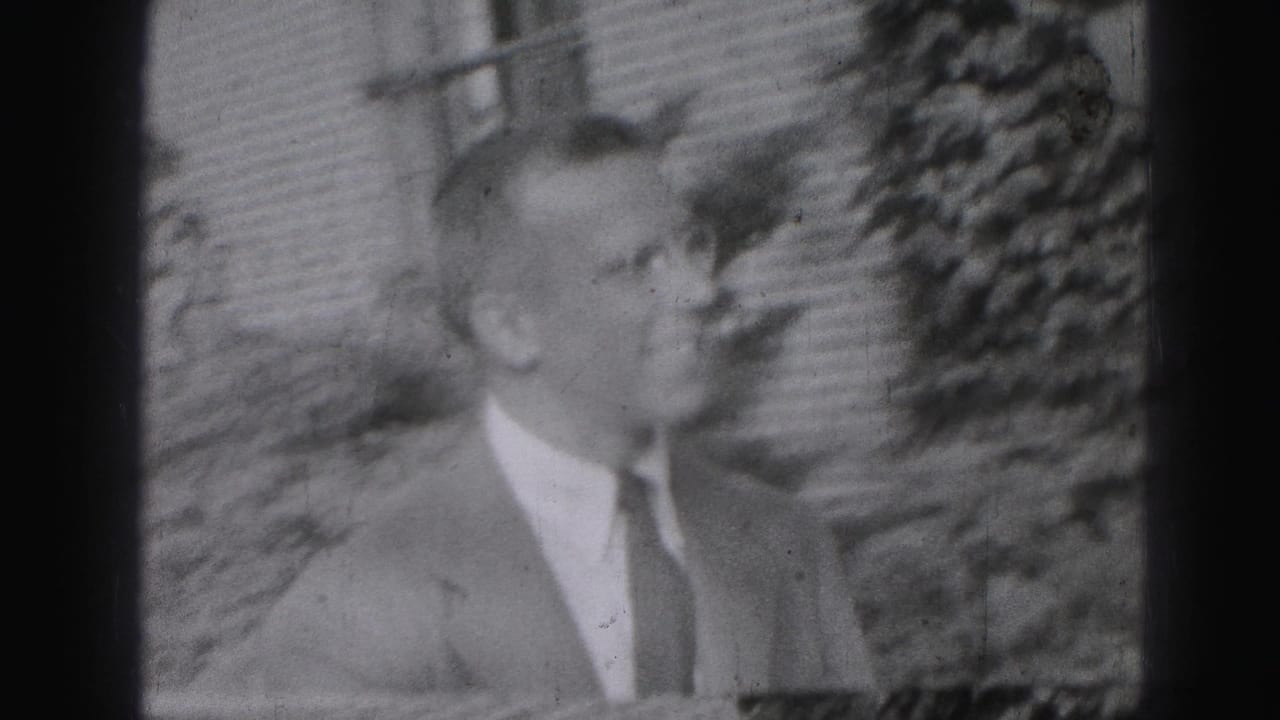 The suited man appears in profile outdoors in the 1975 Harrisburg footage, with a brick facade and foliage in the background.