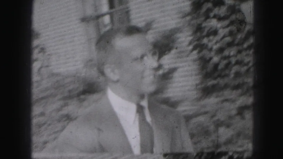 The suited man appears in profile outdoors in the 1975 Harrisburg footage, with a brick facade and foliage in the background.