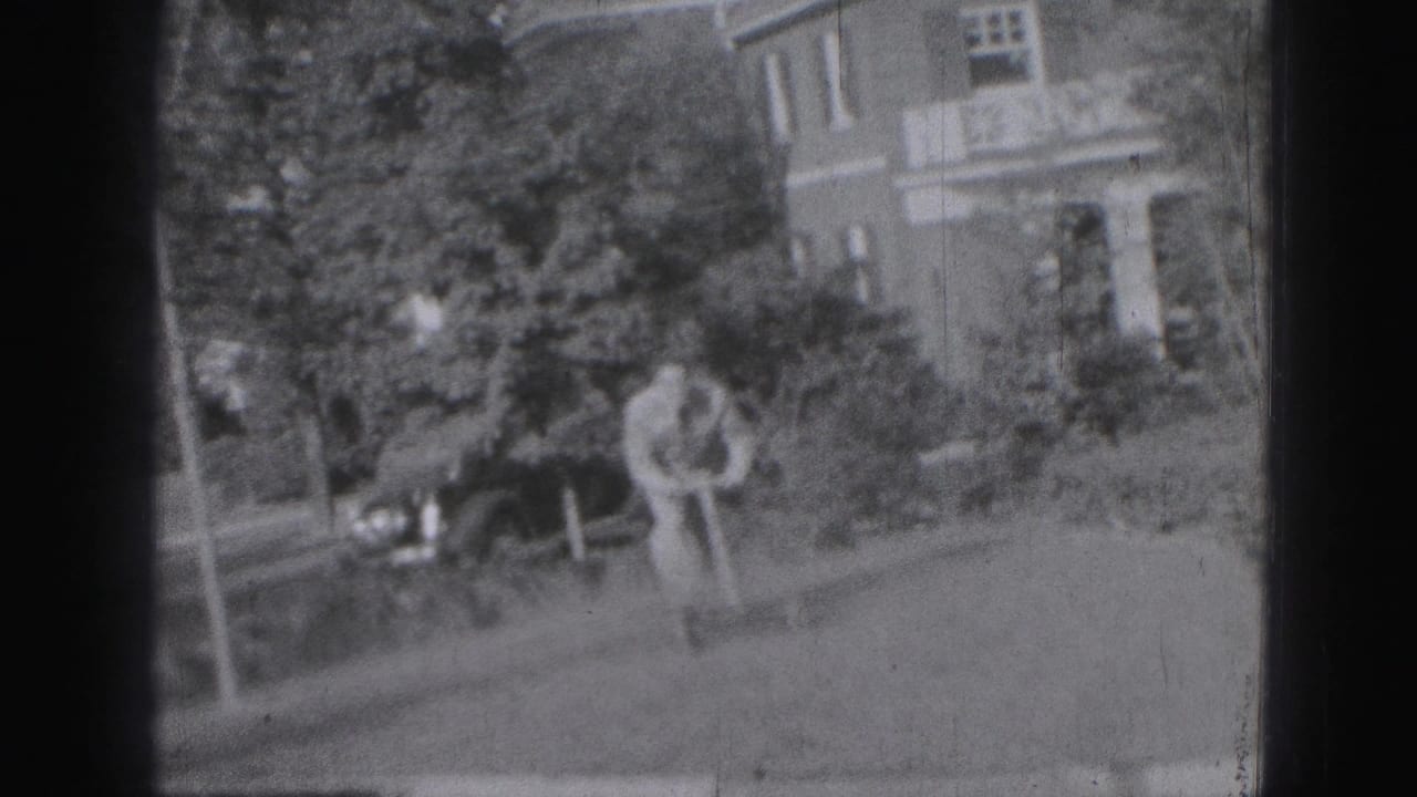 Man pushes a lawnmower across a yard with trees, shrubs, and a brick house behind him.