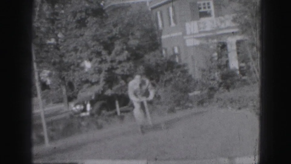 A man mows a grassy yard in Harrisburg, Pennsylvania, with trees, shrubs, and a brick residential facade in the background.