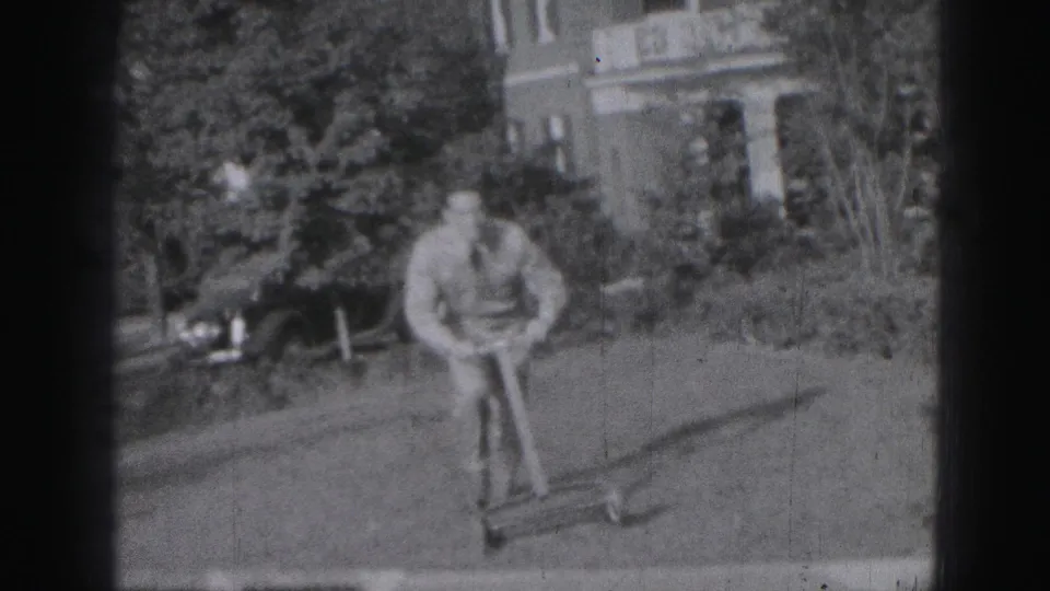 The 1975 home-movie frame shows the man mowing across the yard, with planted shrubs and a brick house visible behind him.