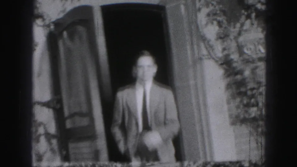 Adult man in a suit and tie appears in a doorway, smiling toward the camera in black-and-white 8mm footage.