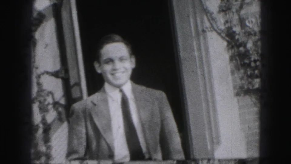 Closer black-and-white home-movie frame of the smiling adult man in formal clothes at the doorway.