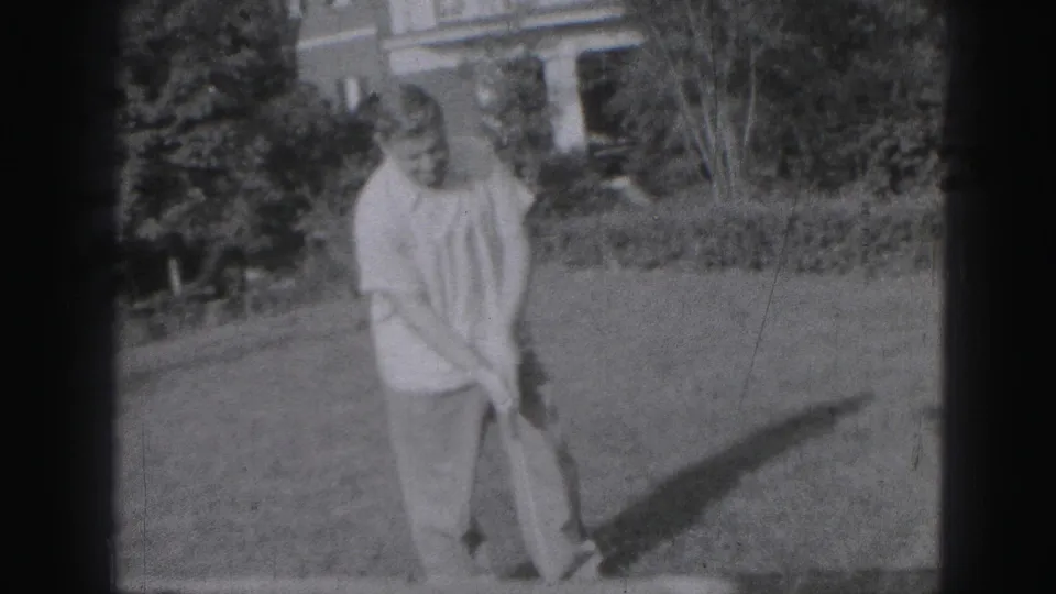 Adult man in a light shirt stands in a Harrisburg yard with a golf club, with shrubs and a house behind him in 1975 home-movie footage.